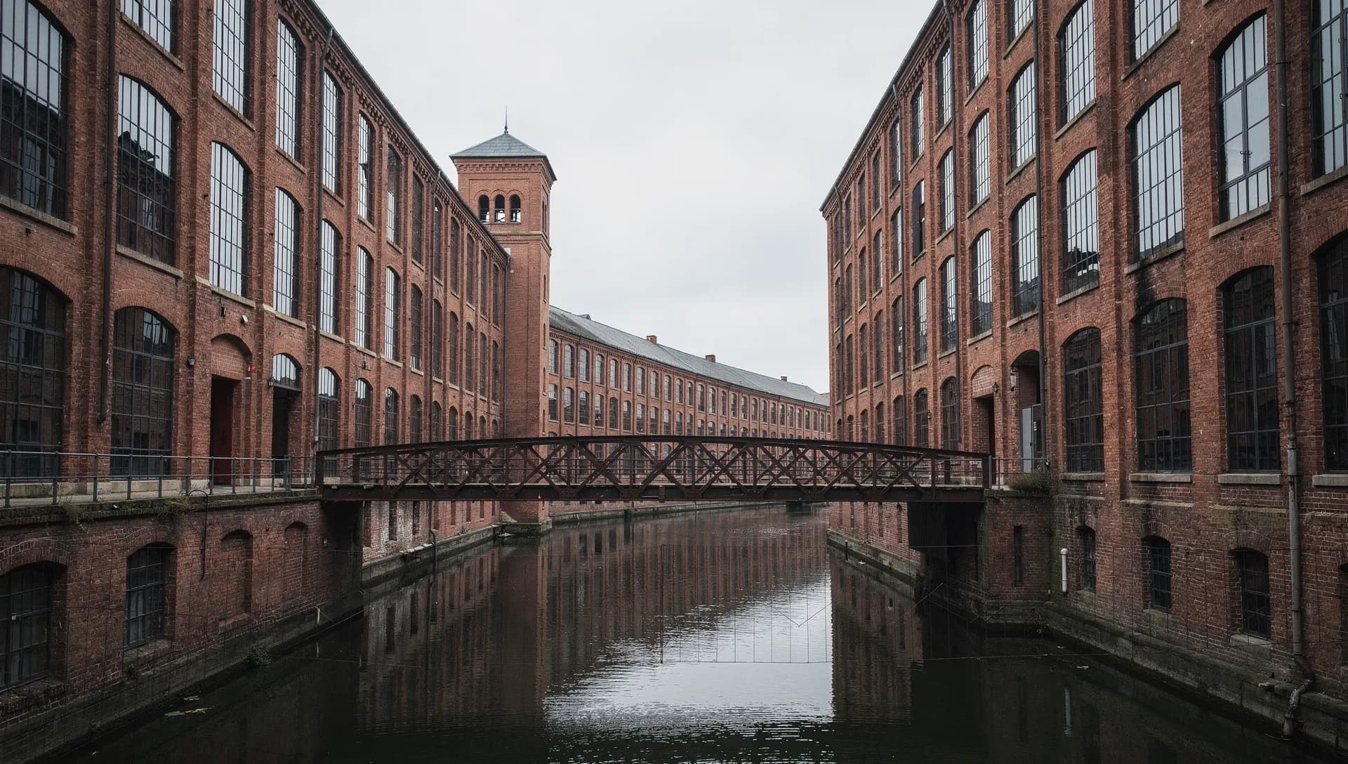 A still canal running between towering red-brick textile mills with rows of tall arched windows, an iron truss footbridge crossing the water and a brick bell tower rising at the center under an overcast sky