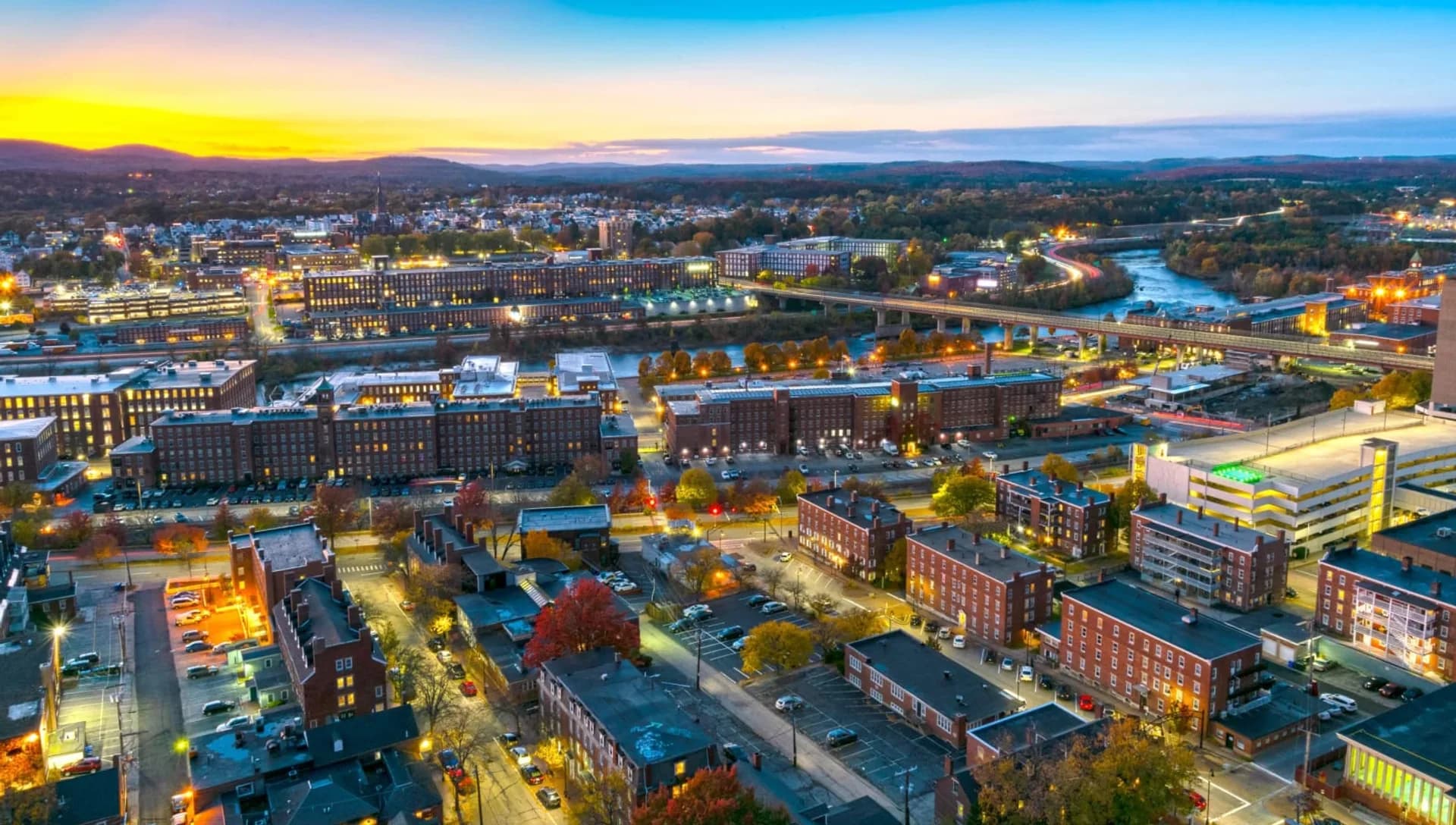 An aerial view at dusk of long brick mill buildings lining both banks of a river, city lights coming on beneath an orange sunset over low hills
