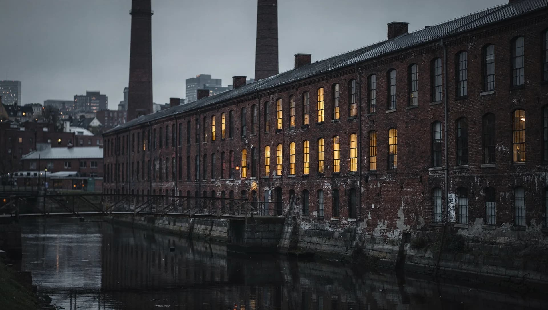 A long brick mill with two tall smokestacks along a canal at dusk, rows of arched windows lit warm, a footbridge crossing the dark water