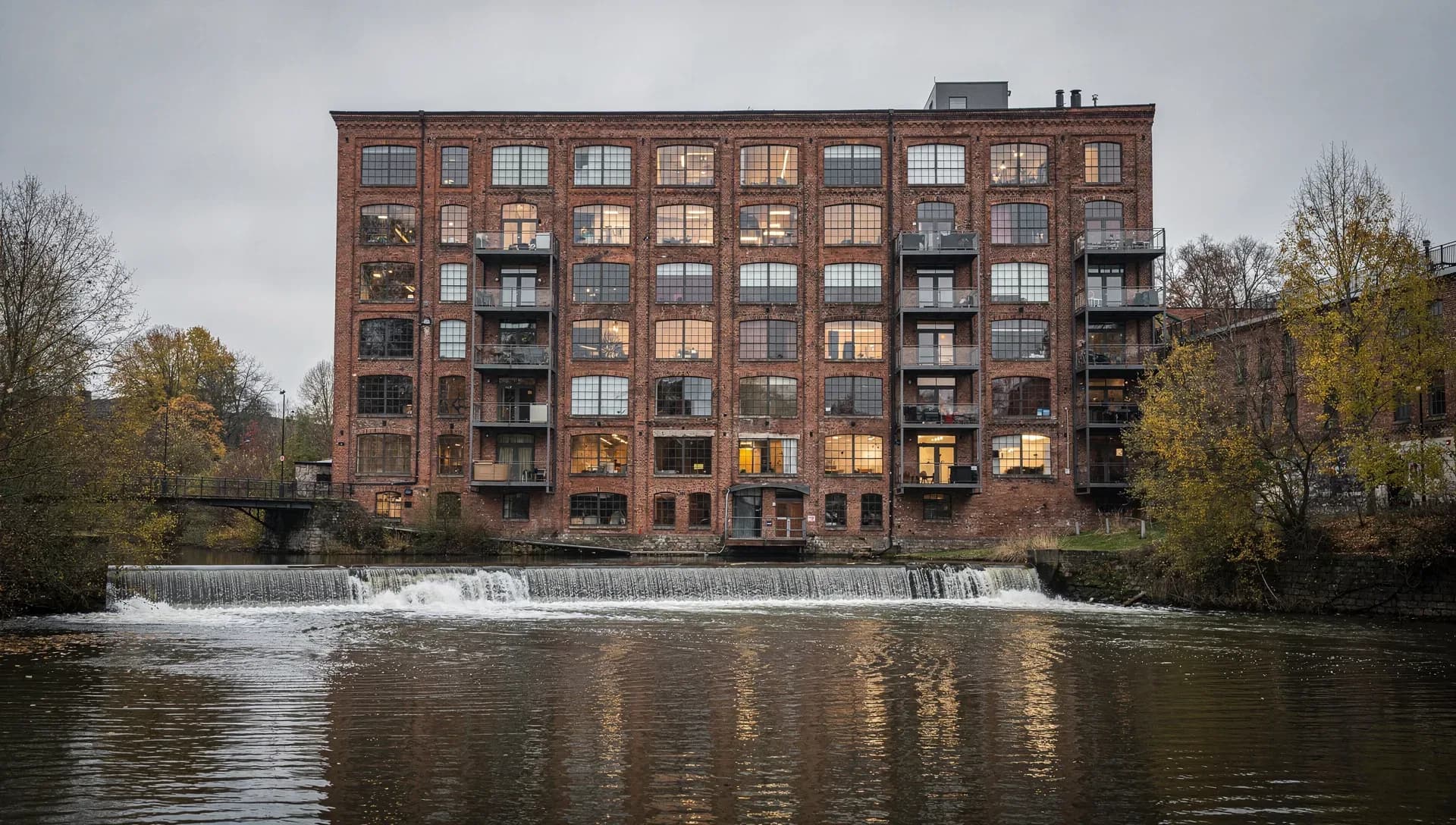 A large restored red-brick riverside mill with rows of tall windows and iron balconies, a low dam spilling white water across the river in front under a grey autumn sky