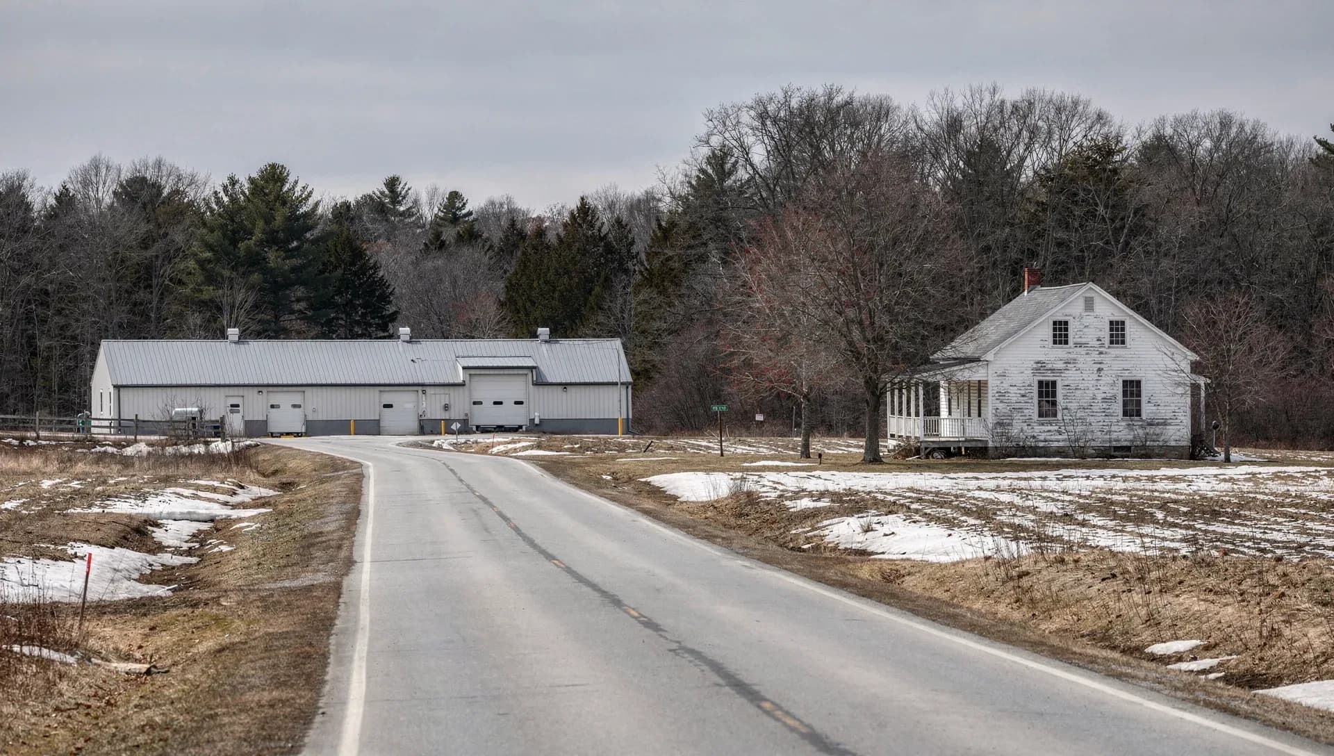 A quiet two-lane country road curving past a long white metal-roofed pole barn with roll-up bay doors on the left and a weathered white farmhouse with a porch on the right, bare trees and evergreens behind patchy melting snow on brown fields under a gray sky