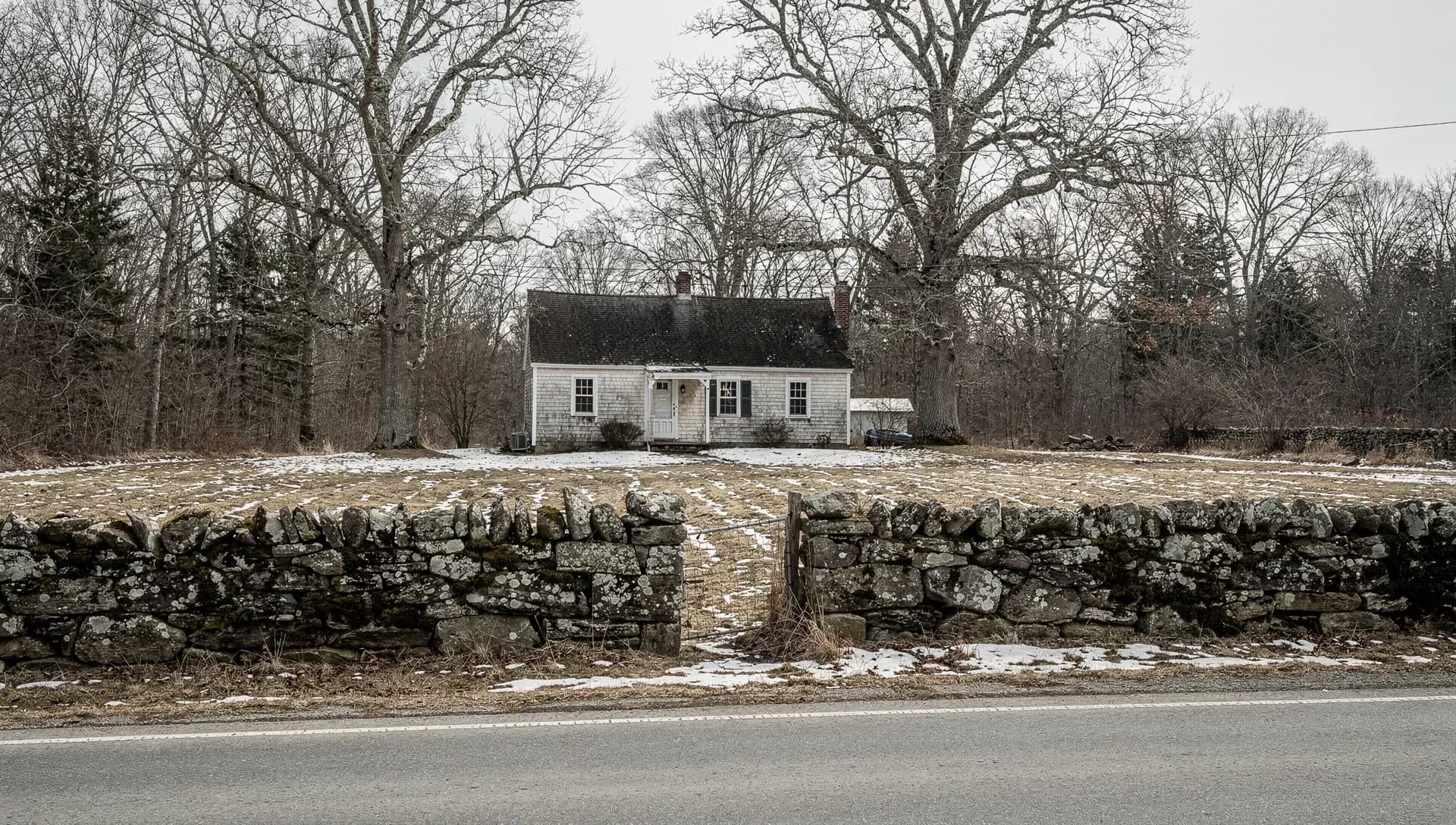 A white Cape-style farmhouse set back behind a low lichen-covered stone wall and a bare winter field, tall leafless oaks flanking it under an overcast sky