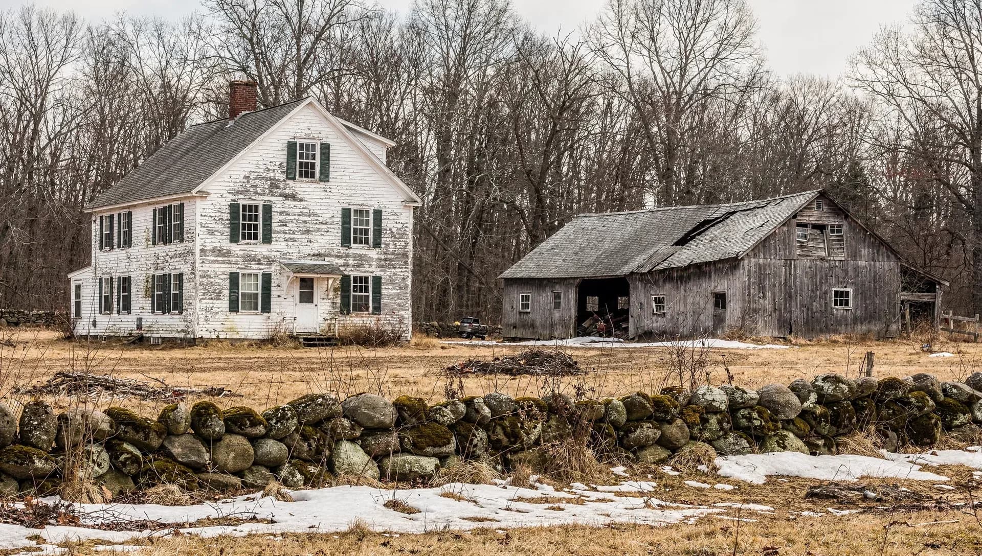 A weathered white farmhouse with green shutters and peeling paint beside a gray weathered barn with an open bay, bare winter woods behind, a moss-covered fieldstone wall across the foreground with dry grass and patches of snow