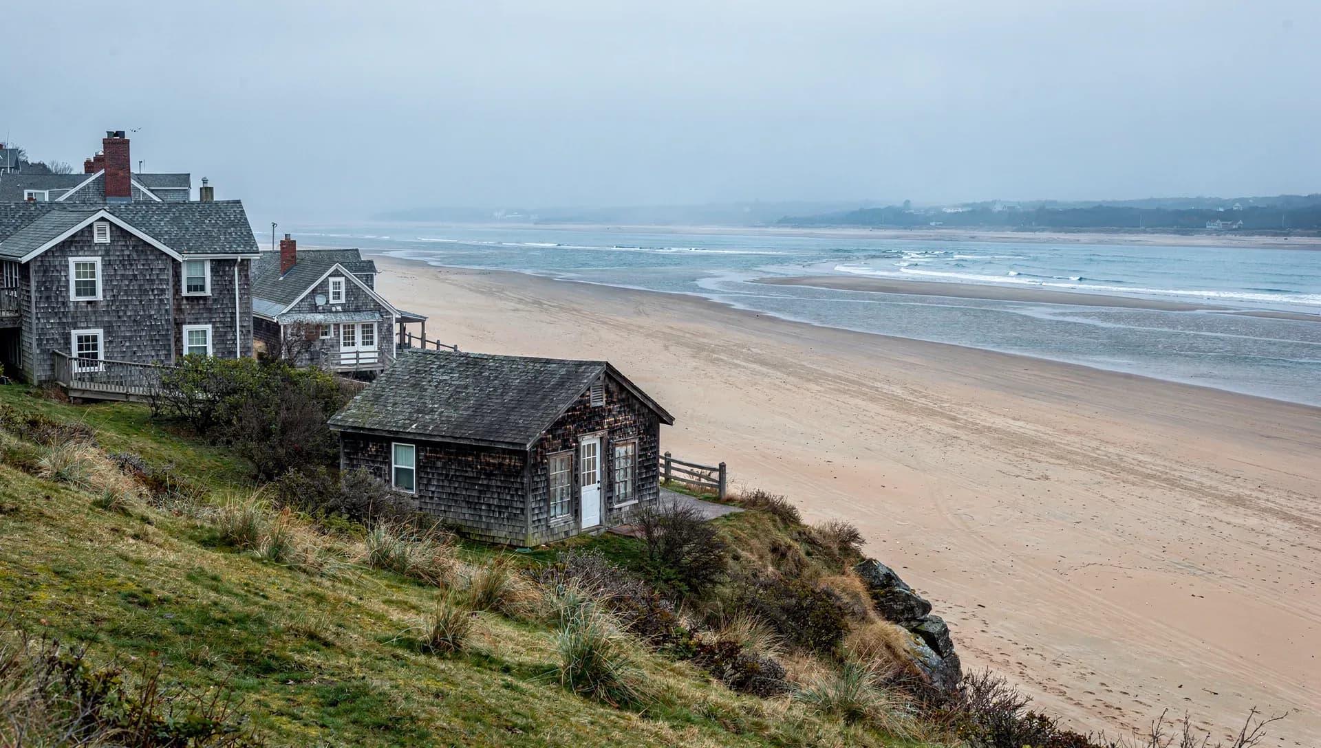 Weathered gray-shingled cottages perched on a grassy dune bluff above a wide, empty sand beach, low surf rolling in under a misty overcast sky with a wooded headland in the distance