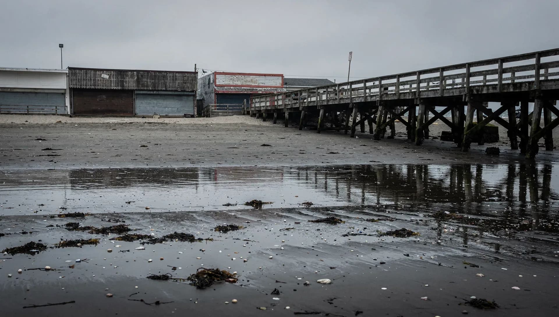 A long weathered wooden pier on pilings stretching over wet low-tide sand strewn with seaweed and shells, shuttered boardwalk buildings behind it under a flat gray overcast sky