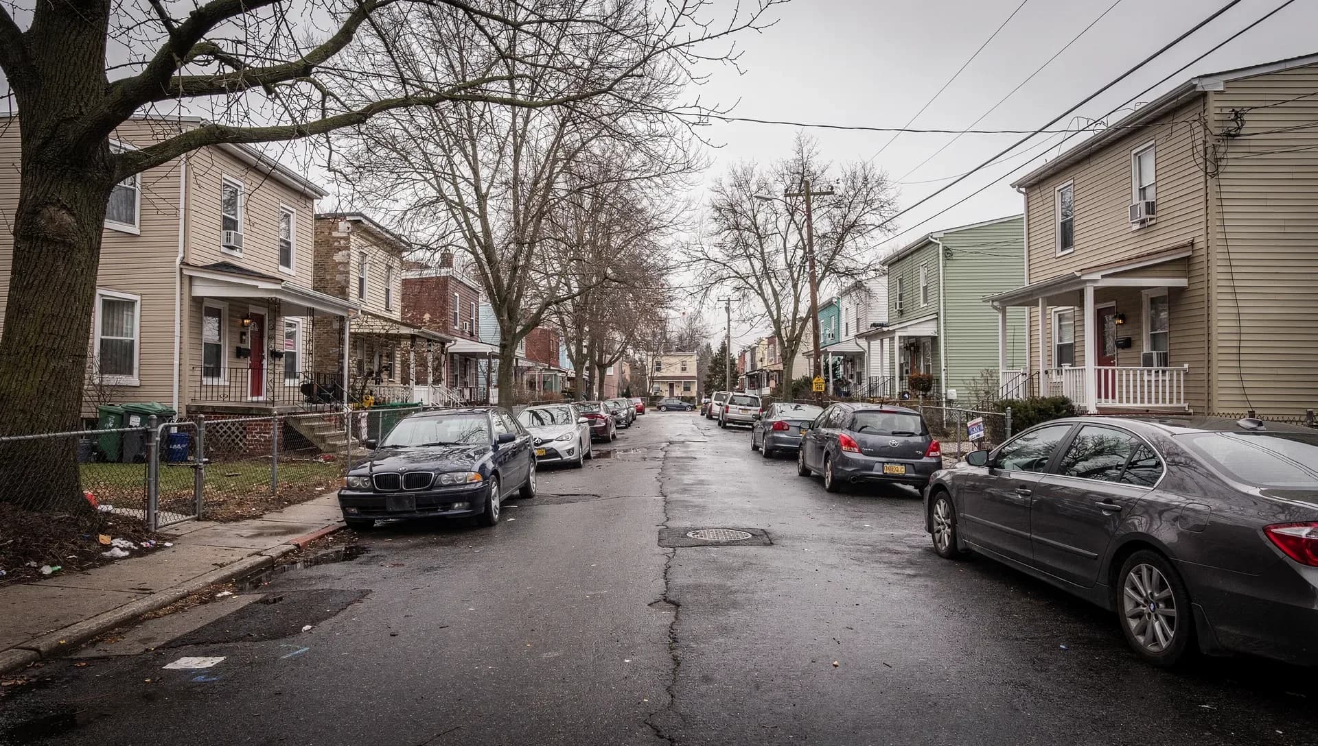 A dense residential side street on an overcast day, tightly packed two-story vinyl-sided and clapboard houses with front porches and chain-link fences, bare trees overhead and cars parked along both wet curbs