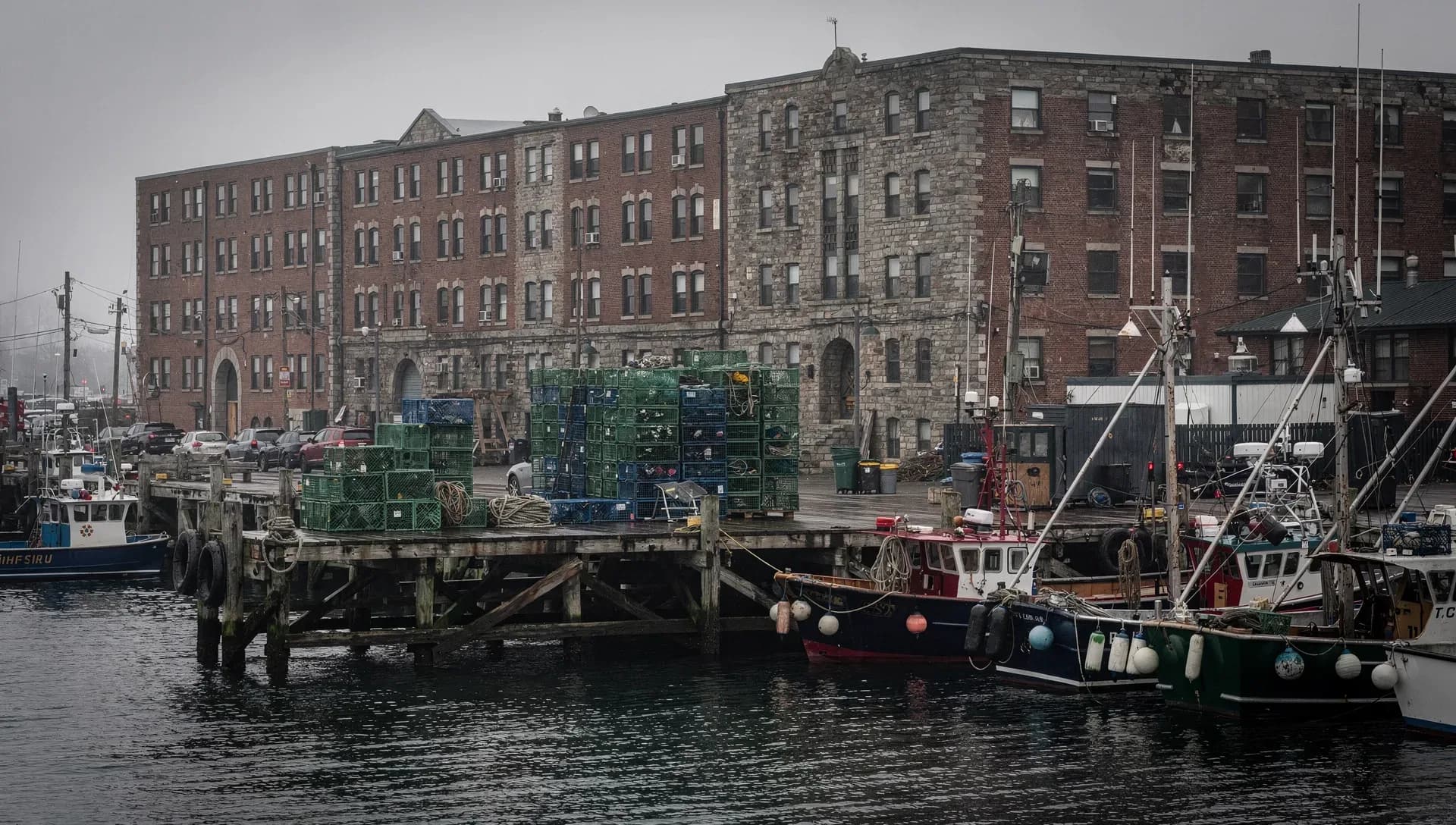 Fishing boats moored along a timber pier stacked with green and blue lobster crates and coiled rope, tall brick and stone waterfront warehouses rising behind under a foggy gray sky