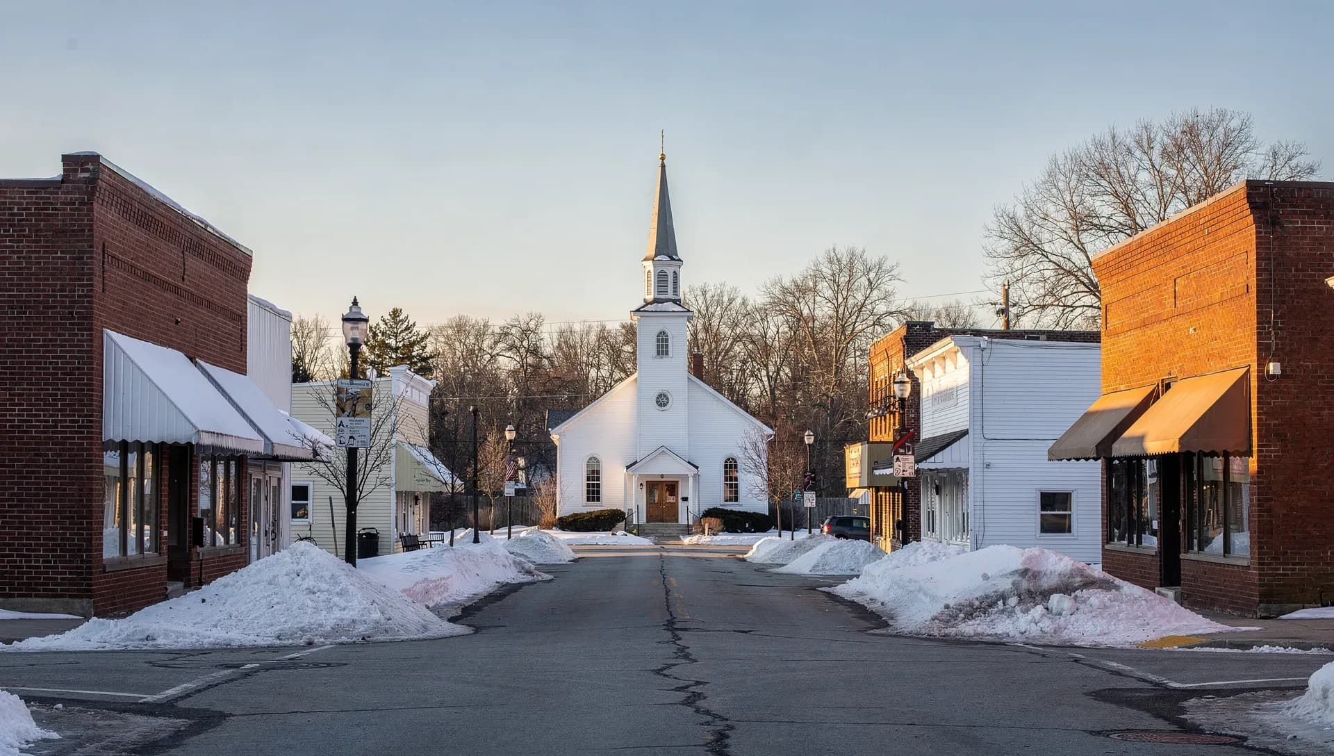 A downtown street at golden hour leading to a white church with a tall steeple, brick commercial buildings with awnings lining both sides above plowed snowbanks and an empty road