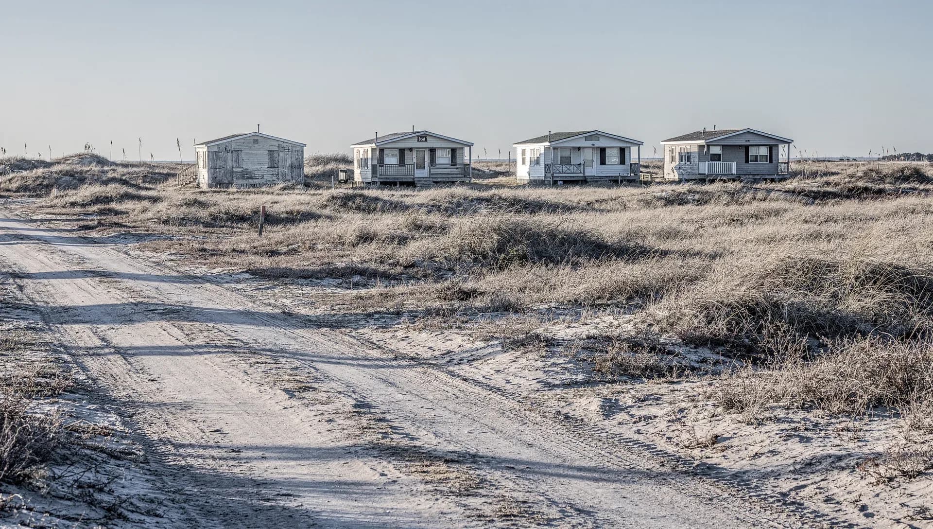 A row of four weathered single-story beach cottages on a low dune, pale dune grass around them, a sandy track with tire ruts crossing the foreground under flat winter coastal light and a wide open sky
