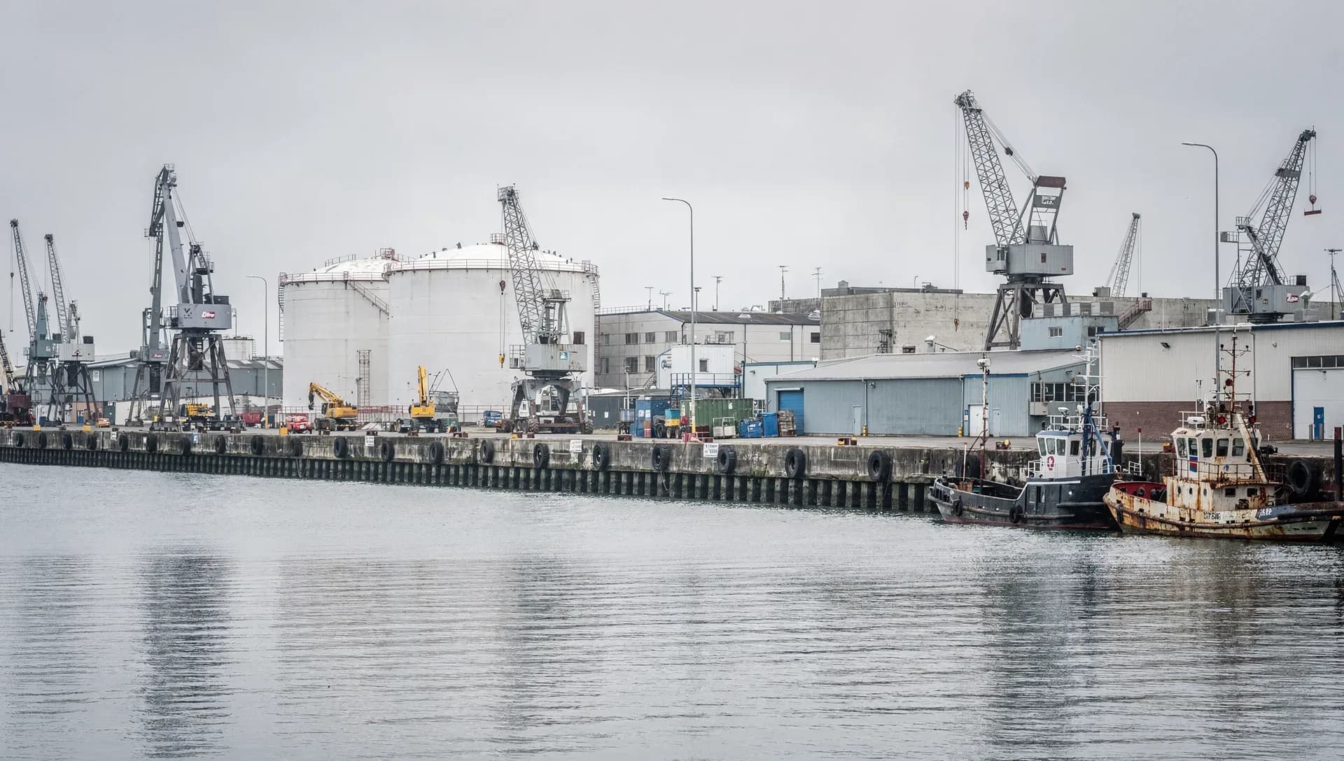An industrial working waterfront with tall white petroleum storage tanks and several dockside cargo cranes behind a concrete pier lined with tire fenders, two tugboats moored at the right under a gray overcast sky