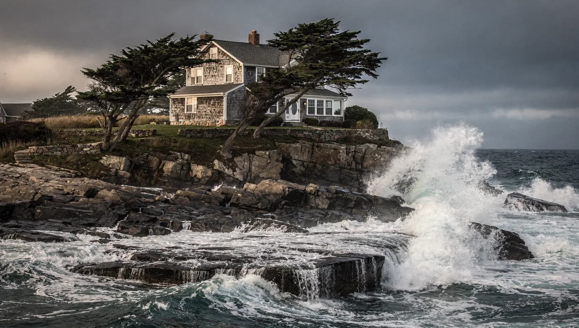 A grey-shingled coastal house on a granite headland framed by wind-bent evergreens, a large wave bursting white against the dark rocks below under a heavy storm sky