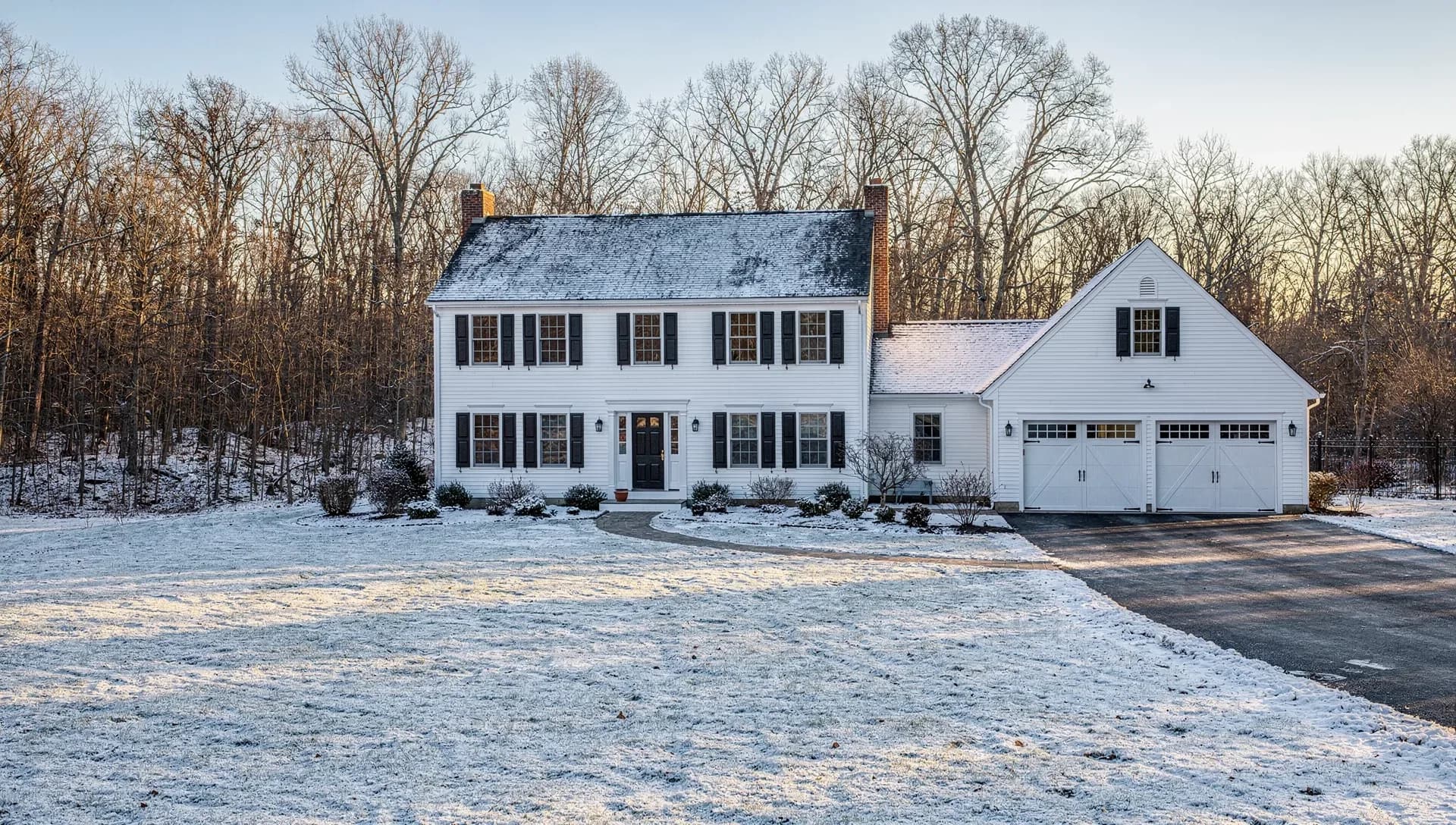 A white two-story colonial house with black shutters and an attached two-car garage on a snow-covered lot at the edge of bare woods