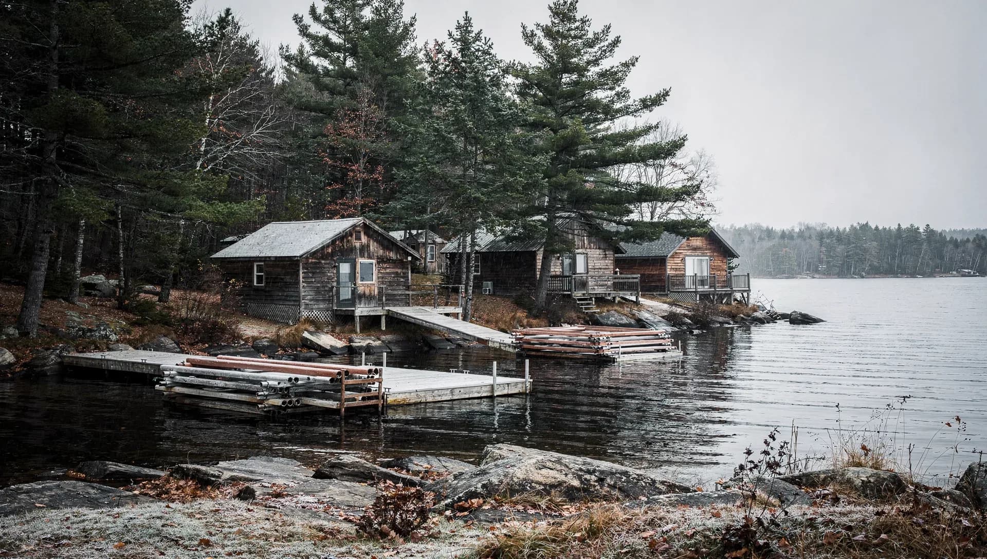 Weathered wood cabins set among tall pines on a rocky lakeshore, sectional wooden docks pulled up with stacked dock hardware for winter, a calm gray lake and forested far shore under an overcast sky with frost on the foreground grass
