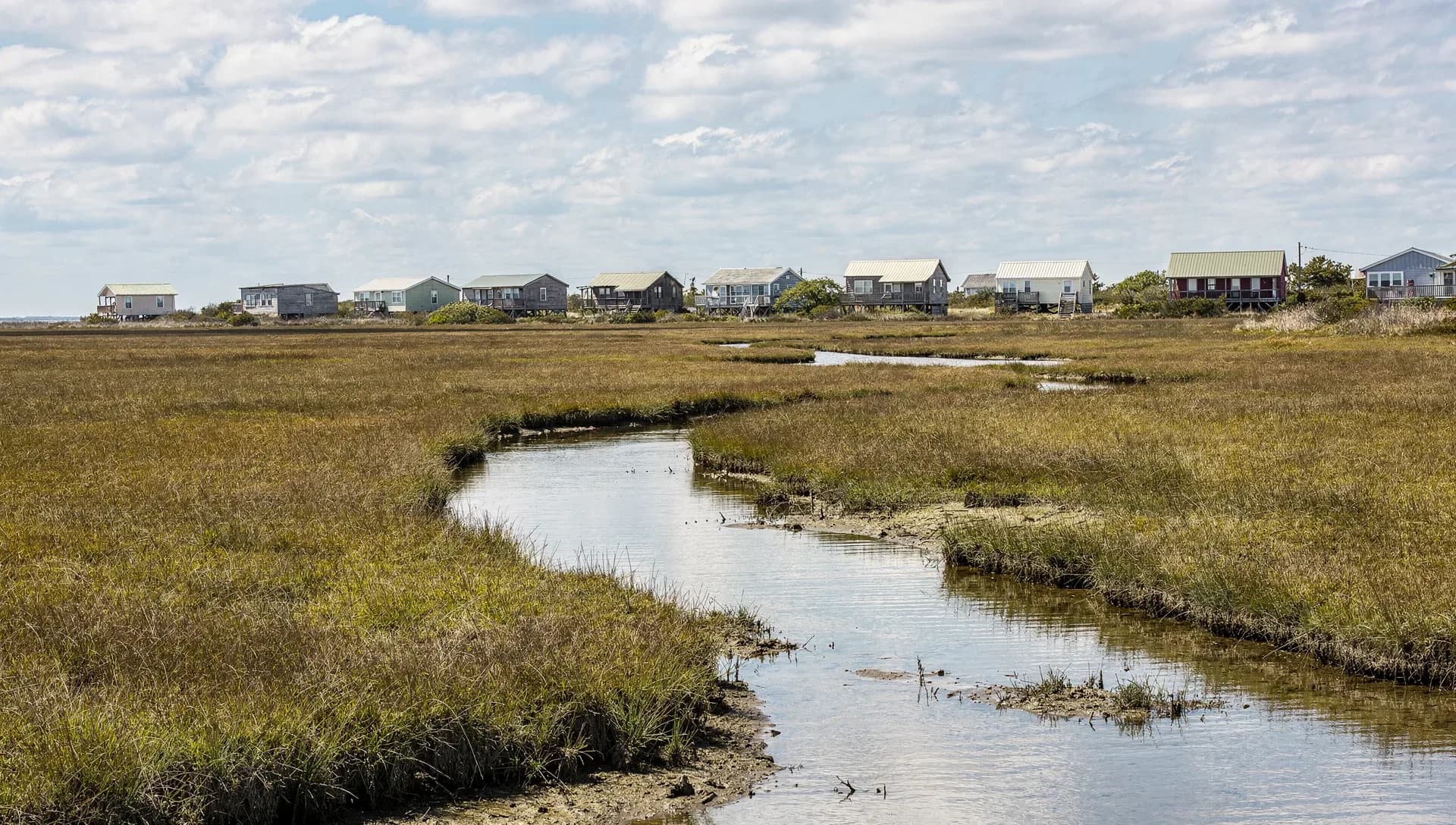 A tidal creek winding through a wide salt marsh of tall golden grass, a line of small single-story beach cottages on low stilts along the horizon under a blue sky scattered with clouds