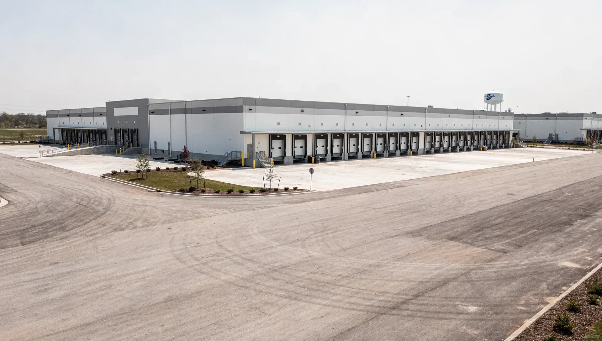 A large modern distribution warehouse with a long row of loading-dock bays and a wide empty concrete truck yard in front, yellow bollards and thin young landscaping trees, a water tower on the hazy horizon