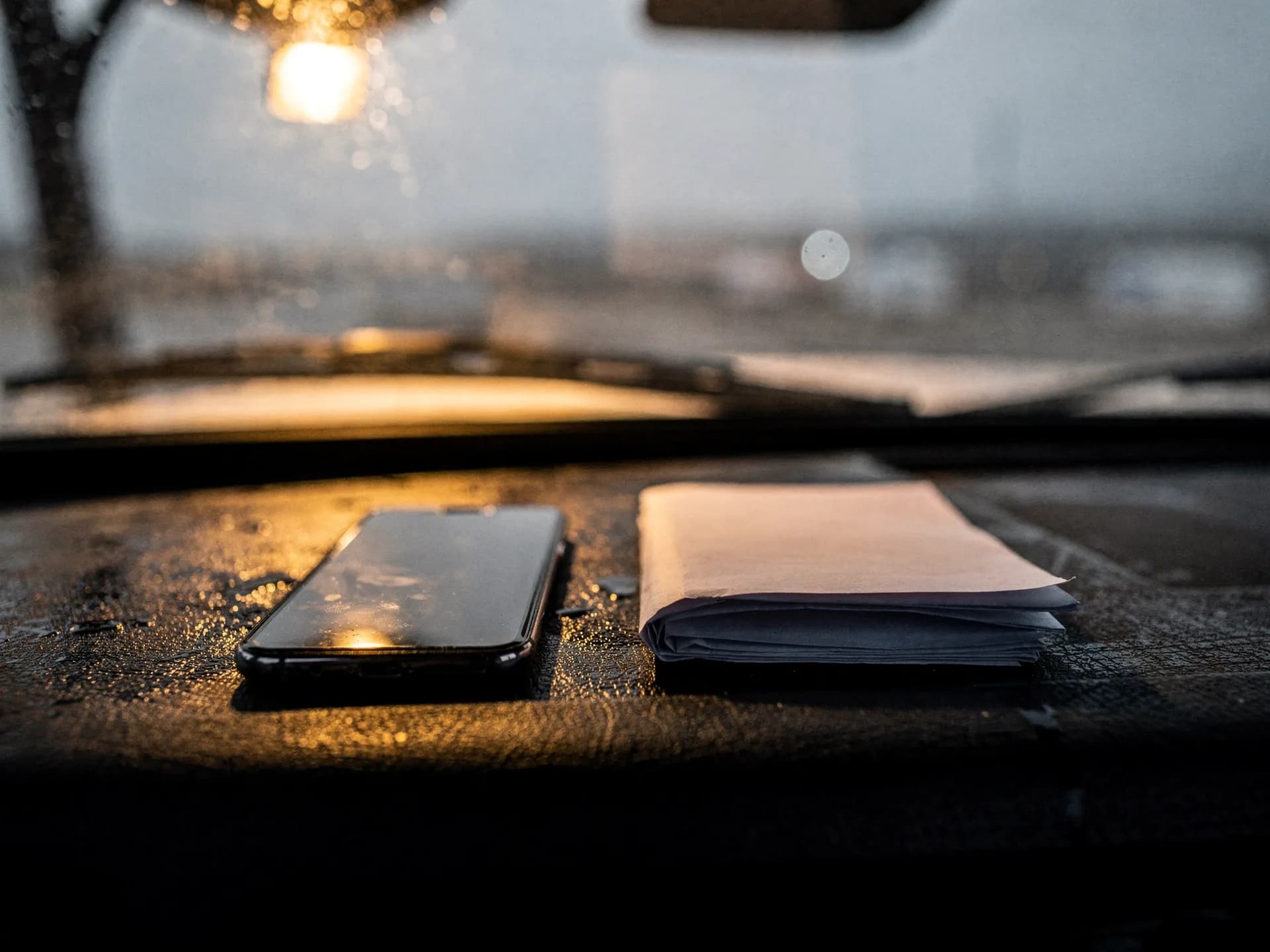 A phone and a folded sheet of paper on a rain-speckled dark dashboard at dusk, a streetlight glowing through the windshield
