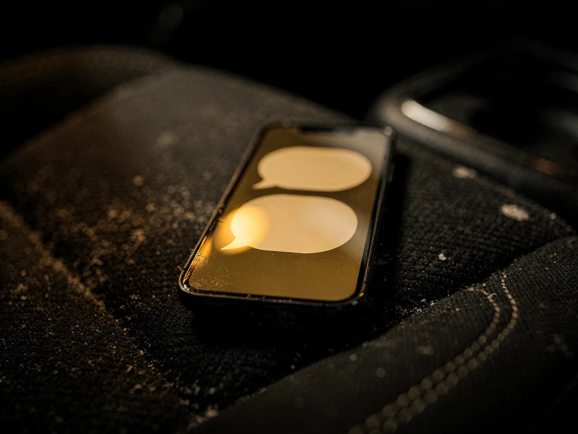 A smartphone lying on a black cloth seat, its screen showing two blank gold speech-bubble shapes