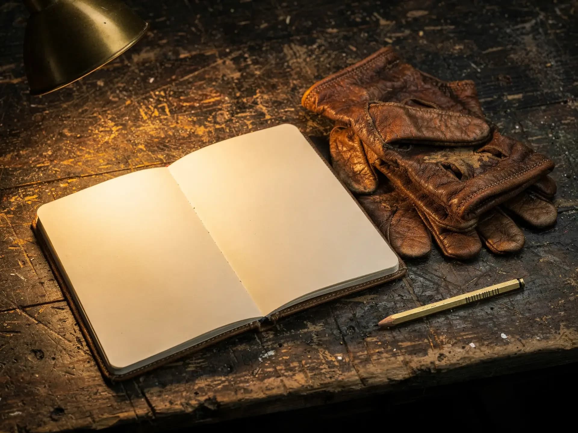 Open blank notebook under a brass lamp on a paint-flecked workbench beside worn leather gloves and a brass pencil
