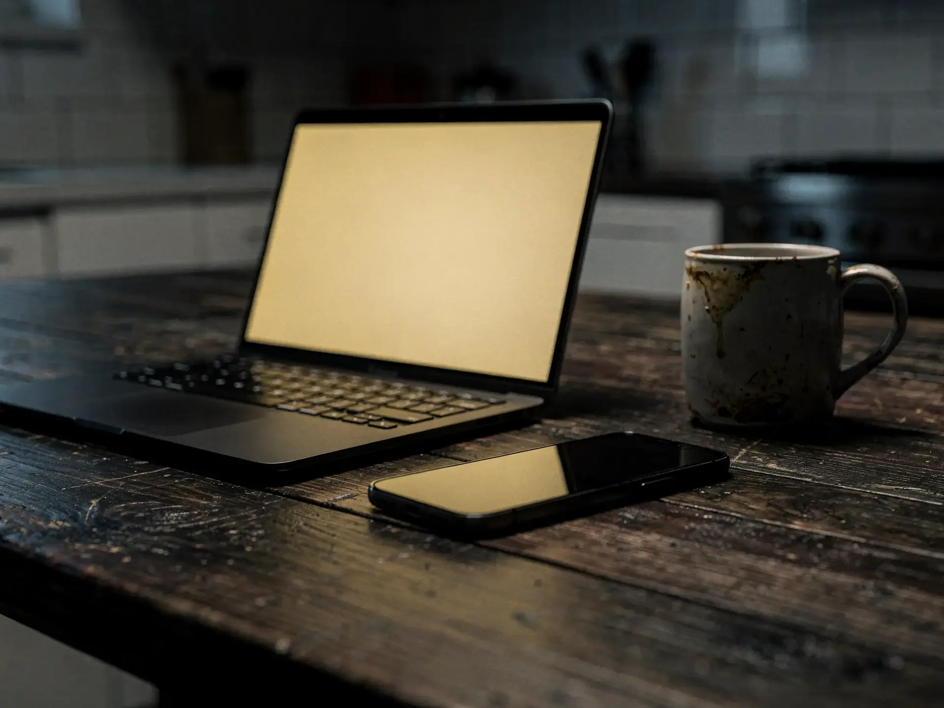 Open laptop and a phone with blank gold-lit screens on a dark wooden kitchen table beside a stained mug