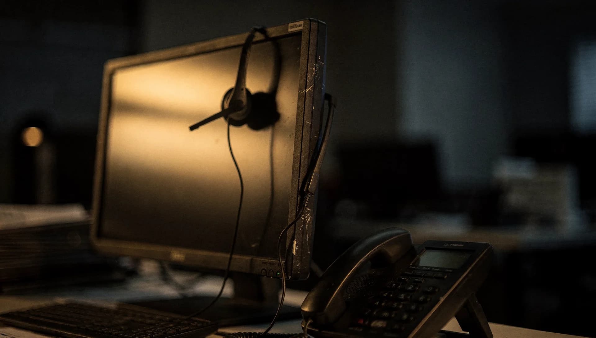 A dark office desk with a headset hung on a monitor and a desk phone in warm side light
